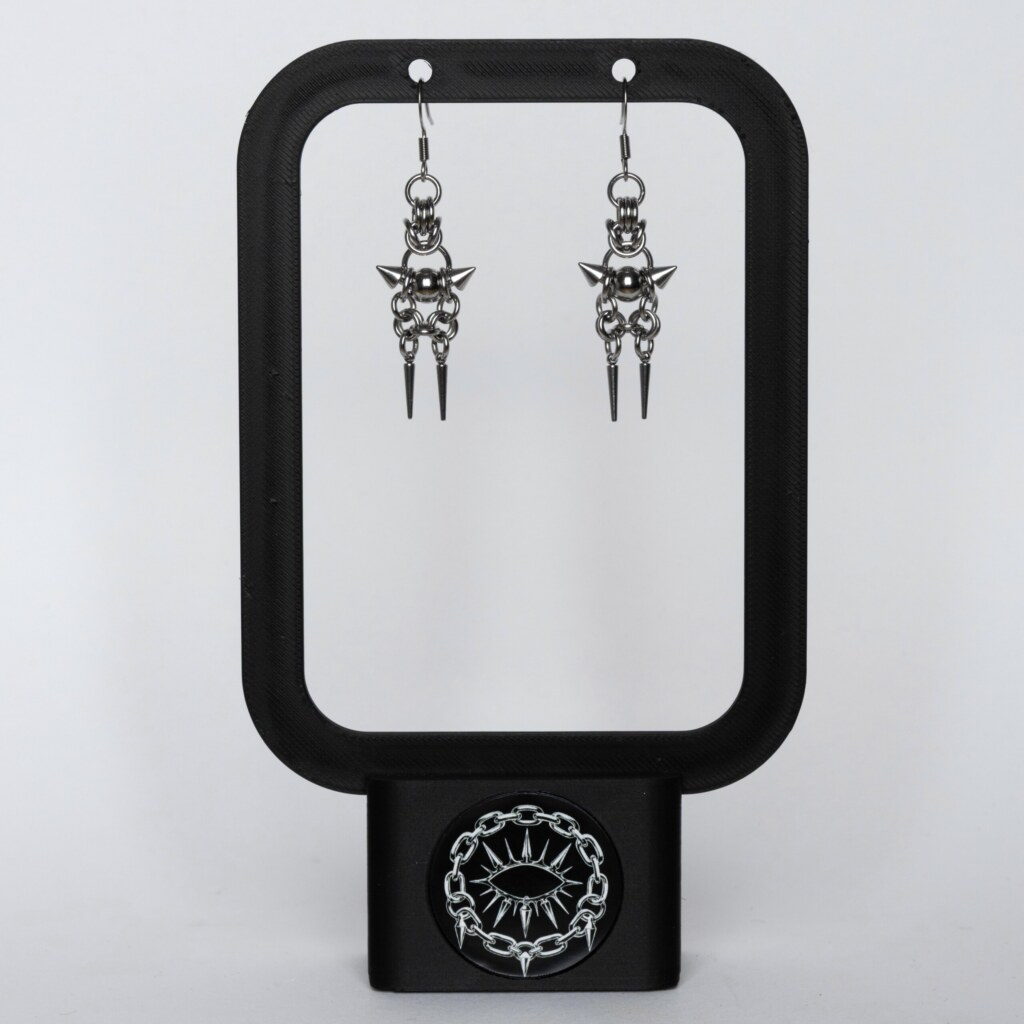 Handmade stainless steel chainmail earrings with a centre bead, side spikes and twin spike drops, displayed on a black earring stand against a light background.