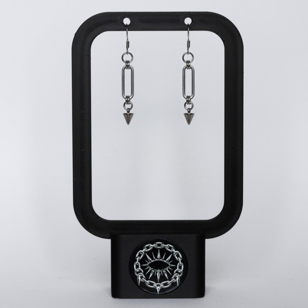 Handmade stainless steel earrings with a large oval chainmail link and small spike drop, displayed on a black earring stand against a light background.