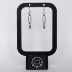 Handmade stainless steel earrings with a large oval chainmail link and small spike drop, displayed on a black earring stand against a light background.