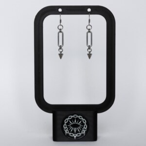 Handmade stainless steel earrings with a large oval chainmail link and small spike drop, displayed on a black earring stand against a light background.