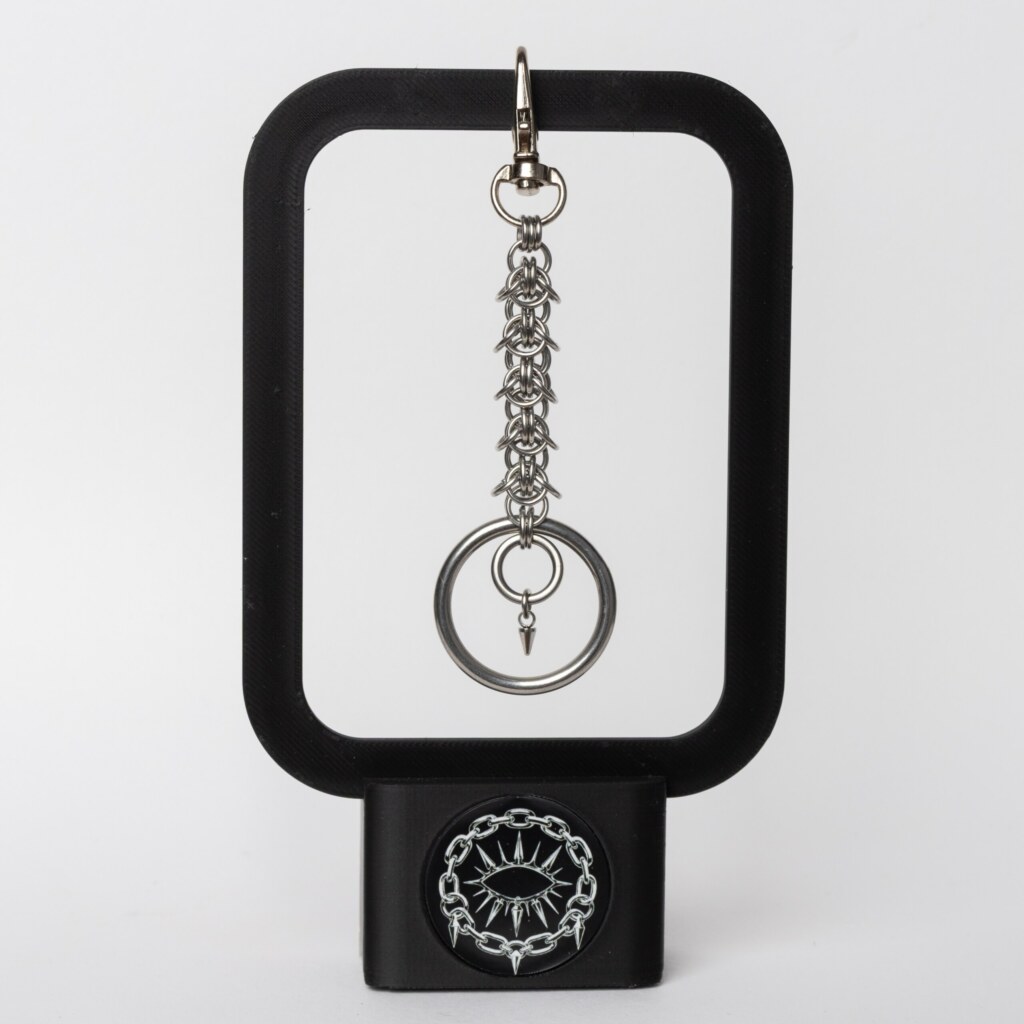 Handmade stainless steel chainmail keychain with swivel clasp, woven chainmail segment, open key ring and small spike drop, displayed on a black stand against a light background.