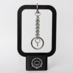 Handmade stainless steel chainmail keychain with swivel clasp, woven chainmail segment, open key ring and small spike drop, displayed on a black stand against a light background.