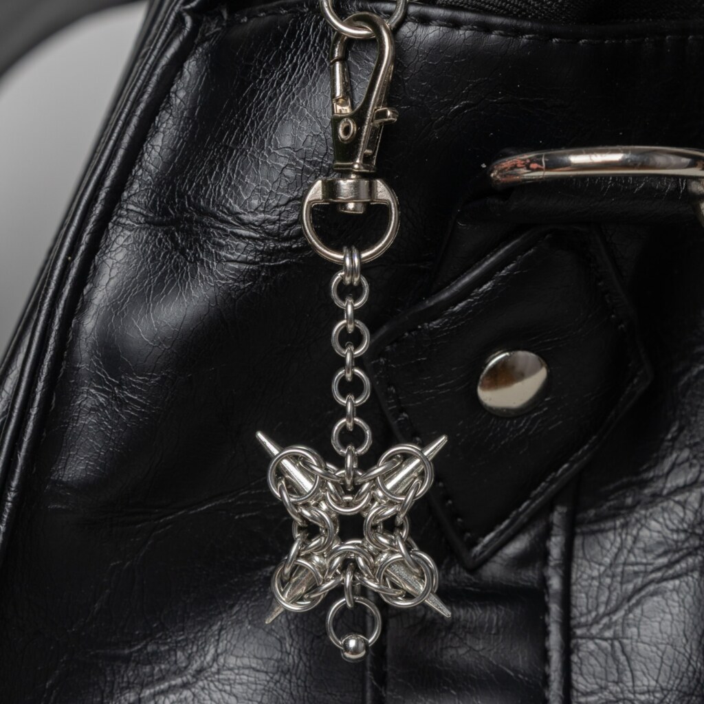 Handmade stainless steel chainmail bag charm with a symmetric four-spike compass design and bead drop, attached to a black bag.