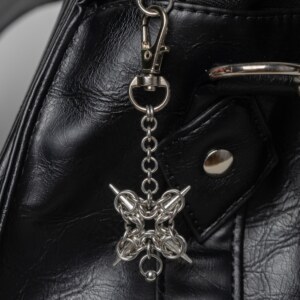Handmade stainless steel chainmail bag charm with a symmetric four-spike compass design and bead drop, attached to a black bag.
