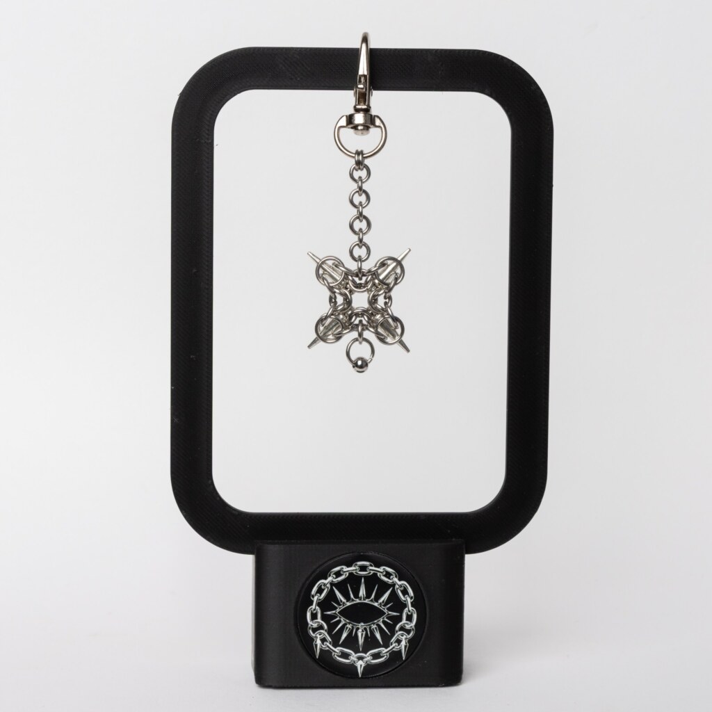 Handmade stainless steel chainmail bag charm with a symmetric four-spike design and bead drop on a swivel clasp, displayed on a black stand against a light background.