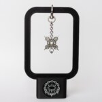 Handmade stainless steel chainmail bag charm with a symmetric four-spike design and bead drop on a swivel clasp, displayed on a black stand against a light background.