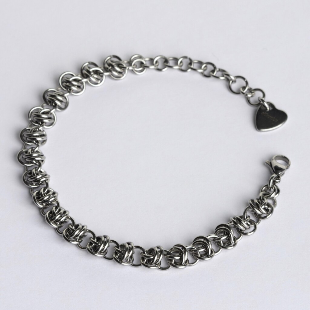 Handmade stainless steel chainmail bracelet with a dense knotted weave pattern, lobster clasp, extension chain and silentchxos heart tag, photographed flat against a light background.