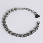 Handmade stainless steel chainmail bracelet with a dense knotted weave pattern, lobster clasp, extension chain and silentchxos heart tag, photographed flat against a light background.