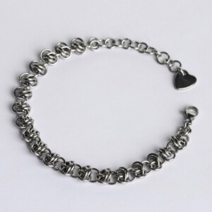 Handmade stainless steel chainmail bracelet with a dense knotted weave pattern, lobster clasp, extension chain and silentchxos heart tag, photographed flat against a light background.