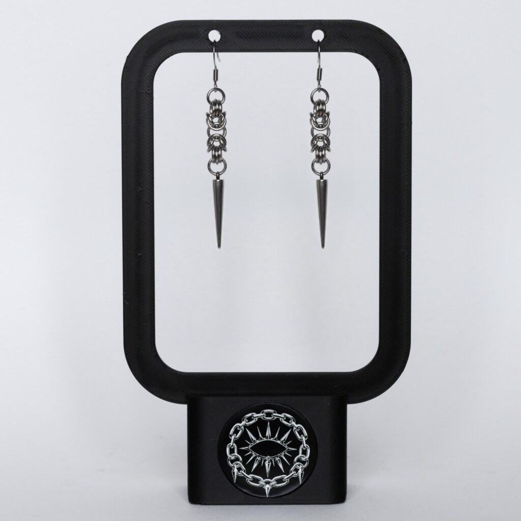 Handmade stainless steel chainmail spike earrings with elongated spike drop, displayed on a black earring stand against a light background.