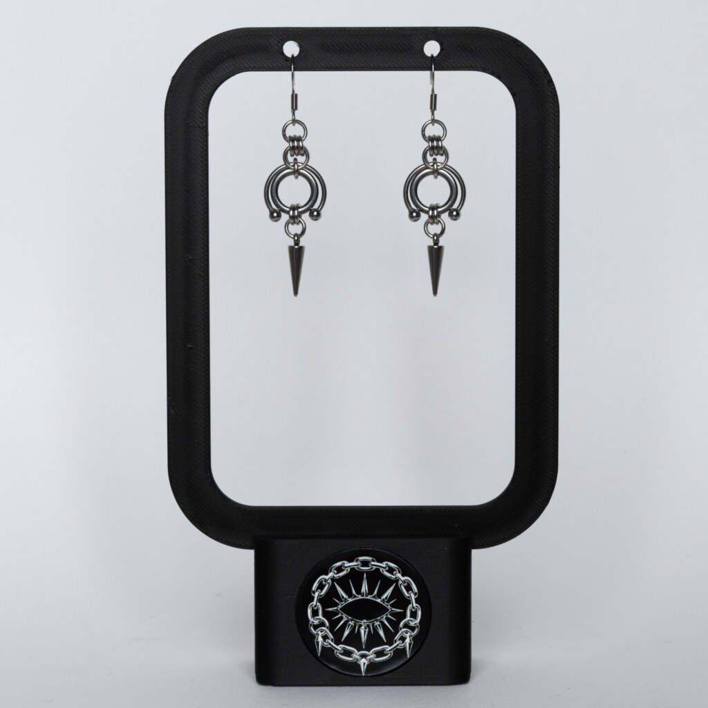 Handmade stainless steel earrings featuring a large open circle ring with bead details and a spike drop, displayed on a black earring stand against a light background.