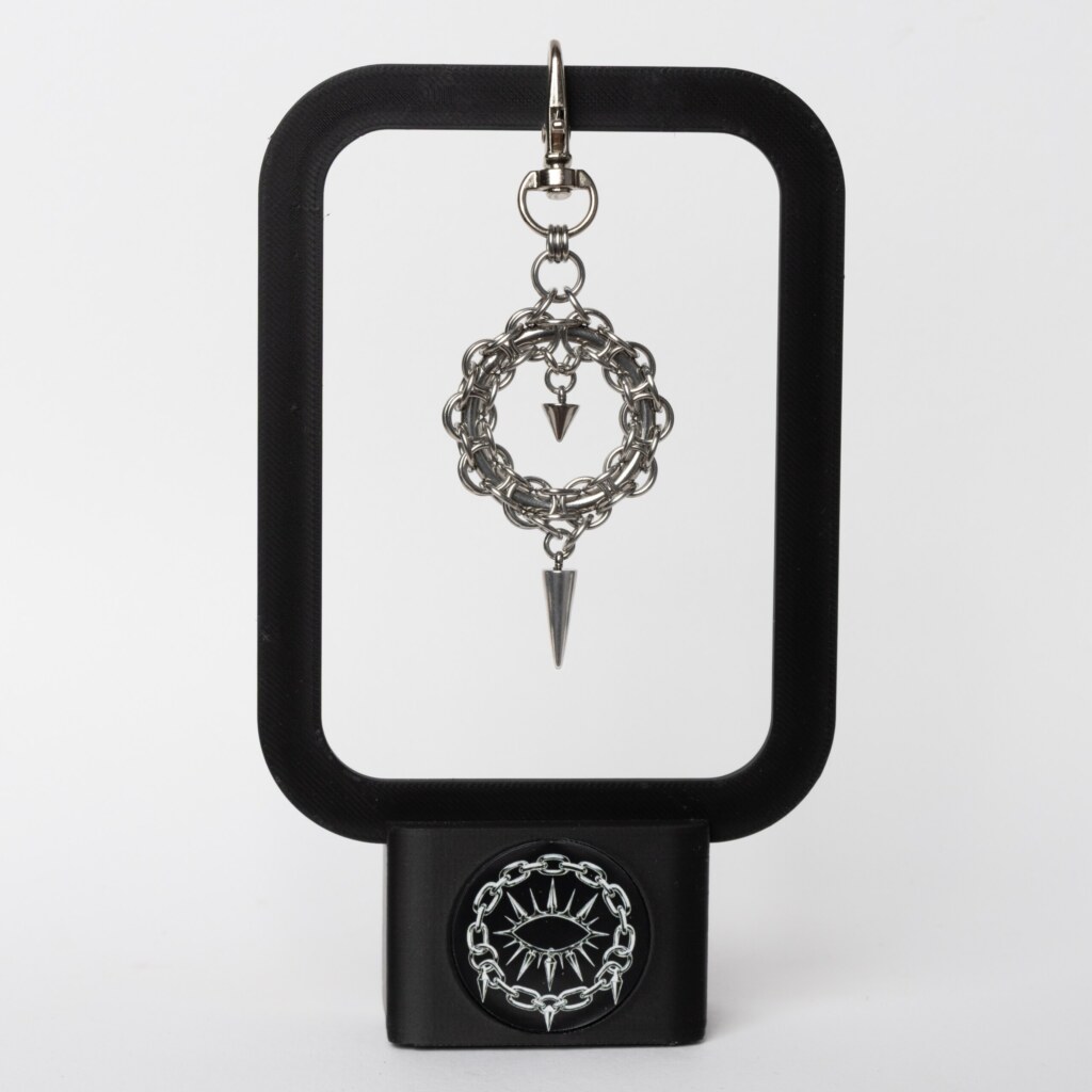 Handmade stainless steel chainmail wreath keychain charm with small inner spike and elongated spike drop on a swivel clasp, displayed on a black stand against a light background.