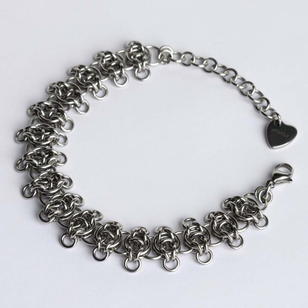 Handmade stainless steel chainmail bracelet with a textured vertebra-inspired weave and small extending rings, lobster clasp, extension chain and silentchxos heart tag, photographed flat against a light background.