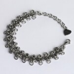 Handmade stainless steel chainmail bracelet with a textured vertebra-inspired weave and small extending rings, lobster clasp, extension chain and silentchxos heart tag, photographed flat against a light background.