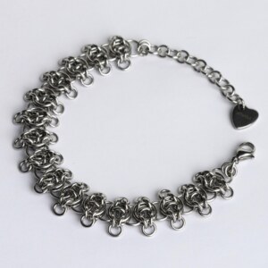 Handmade stainless steel chainmail bracelet with a textured vertebra-inspired weave and small extending rings, lobster clasp, extension chain and silentchxos heart tag, photographed flat against a light background.