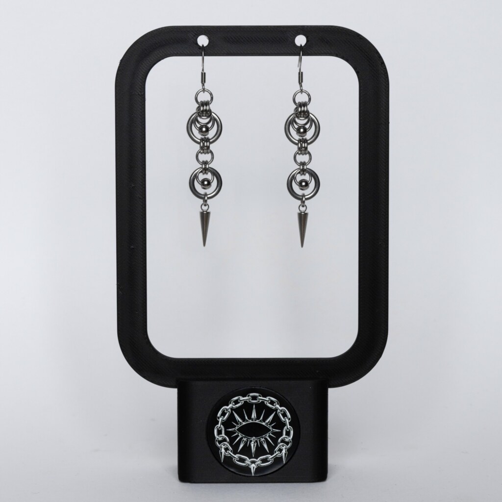 Handmade stainless steel chainmail earrings with repeating ring and bead links leading to a long spike drop, displayed on a black earring stand against a light background.