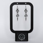 Handmade stainless steel chainmail earrings with repeating ring and bead links leading to a long spike drop, displayed on a black earring stand against a light background.