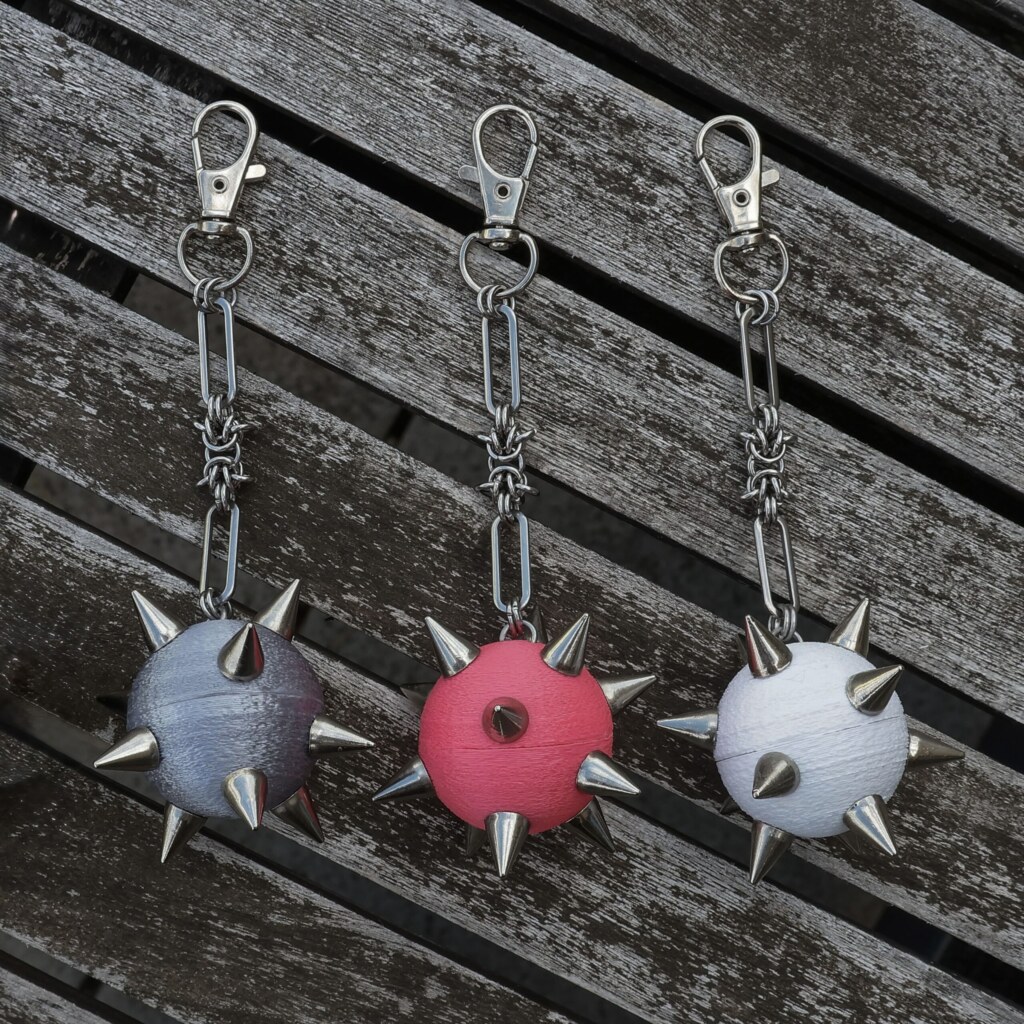 Handmade bag charm with a spiked ball pendant and stainless steel paperclip chain in different colors, lying on an table.